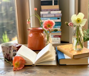 Image is of an open book next to two other books, with small jars of poppies around, a coffee and a stack books in the background.