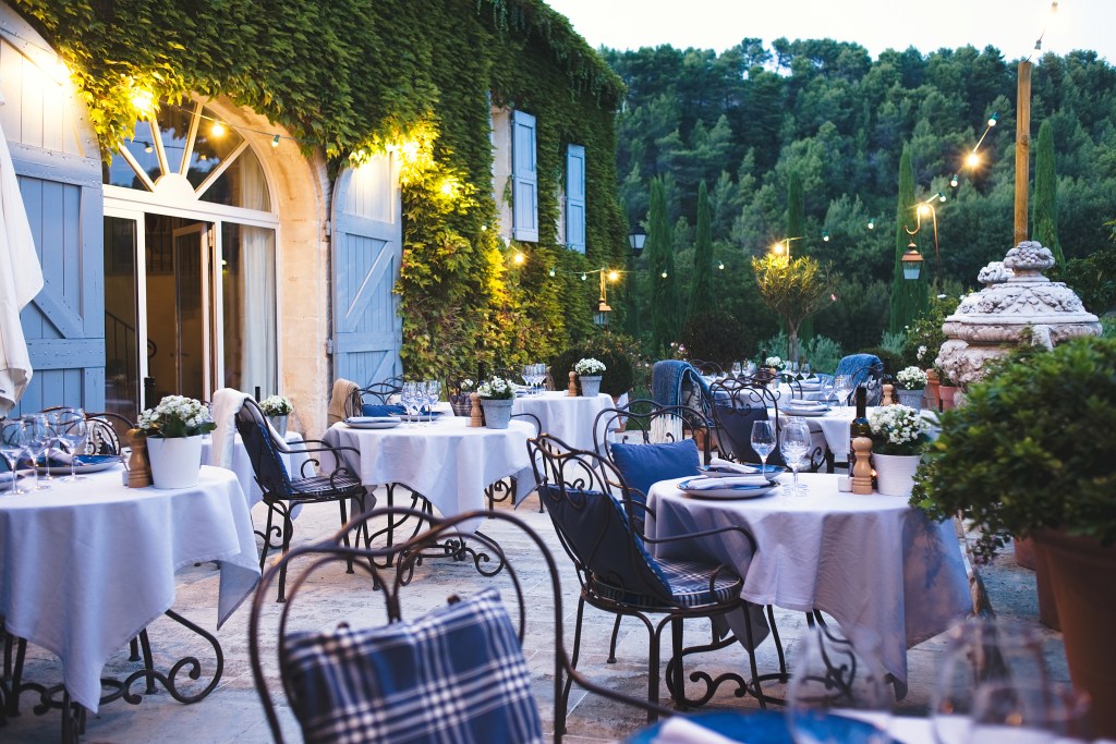 Outdoor dining area of a restaurant with elegant tables set for guests, surrounded by greenery and soft lighting.