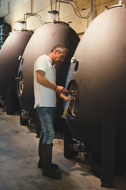 A man in a white shirt and boots inspects a wine fermentation tank in a winery, focusing on the tank's opening as he takes a sample.