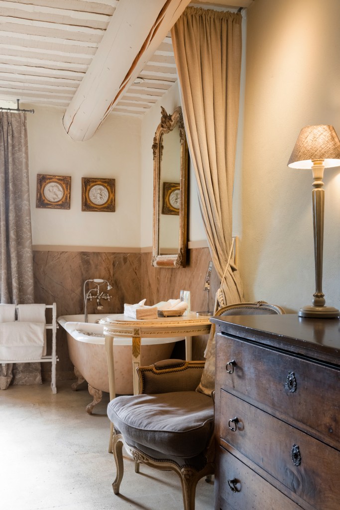 A stylish bathroom featuring a freestanding bathtub, vintage decor, a wooden dresser, and elegant curtains. The space includes a mirror and decorative wall art, displaying a warm and inviting atmosphere.