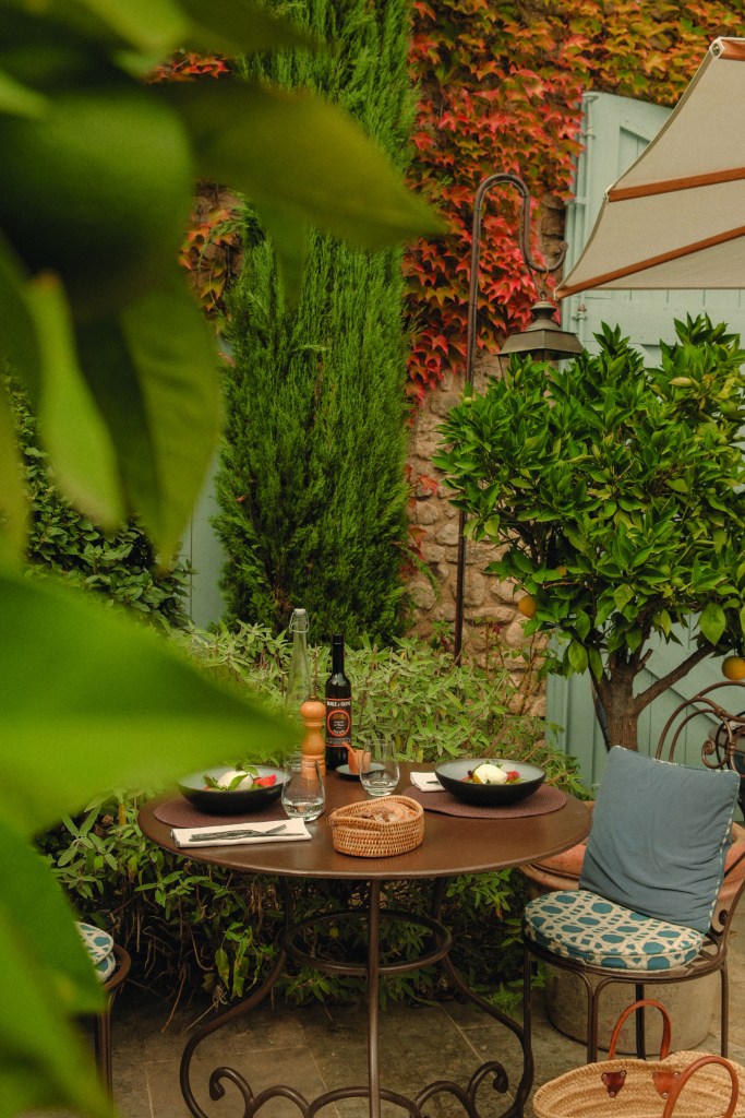 A charming outdoor dining setup featuring a round table with two plates of food, glasses, and a bottle of wine, surrounded by lush green plants and colorful foliage.