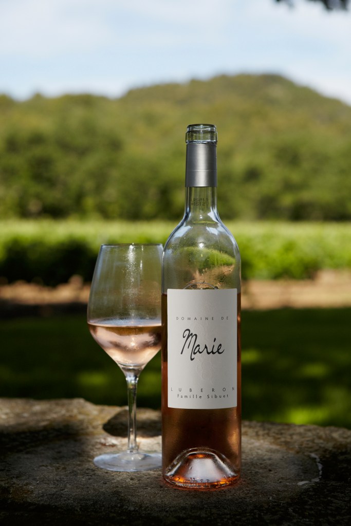 A bottle of Domaine de Marie Luberon rosé wine next to a glass filled with the same wine, set against a blurred background of green vineyards and hills.