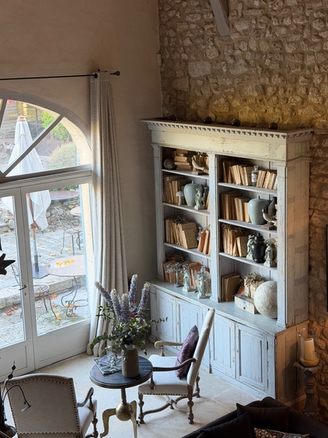 A cozy living space featuring a light blue bookshelf filled with books and decorative ceramic items, next to a small round table with a vase of flowers. Large windows with curtains let in natural light, revealing a glimpse of an outdoor terrace.