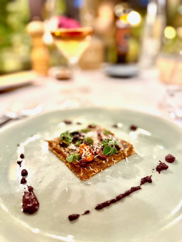 A plated gourmet dish featuring a square pasta layer topped with vegetables, garnished with herbs and a drizzle of sauce on a green plate.