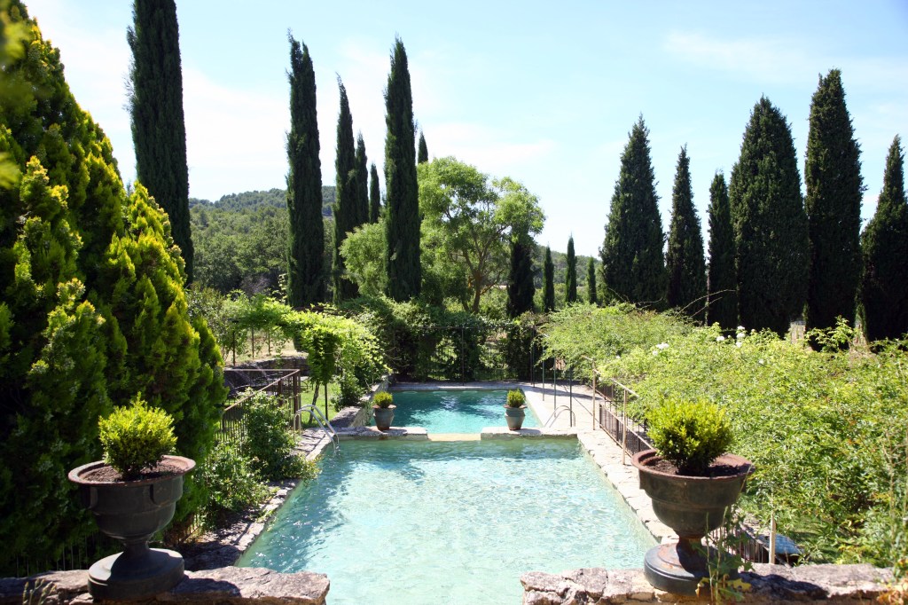 A serene outdoor garden featuring a clear swimming pool surrounded by tall cypress trees, lush greenery, and ornamental planters.