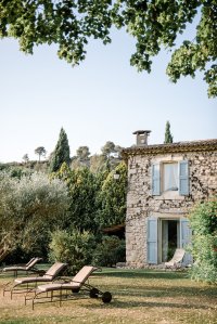 A rustic stone house with blue shutters surrounded by lush greenery, featuring several lounge chairs on a grassy area.